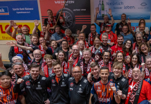 ASC Grünwettersbach setzt Ausrufezeichen zum TTBL-Saisonende in Grenzau Large group of athletes and supporters wearing red and black scarves posing together on a gym floor for a group photo at a sports event.