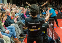 Landesderby zum Heimauftakt in der Tischtennis Bundesliga Back view of a table tennis player in a black jersey with UWE and sponsor logos, arms raised toward a cheering crowd along the red court edge.